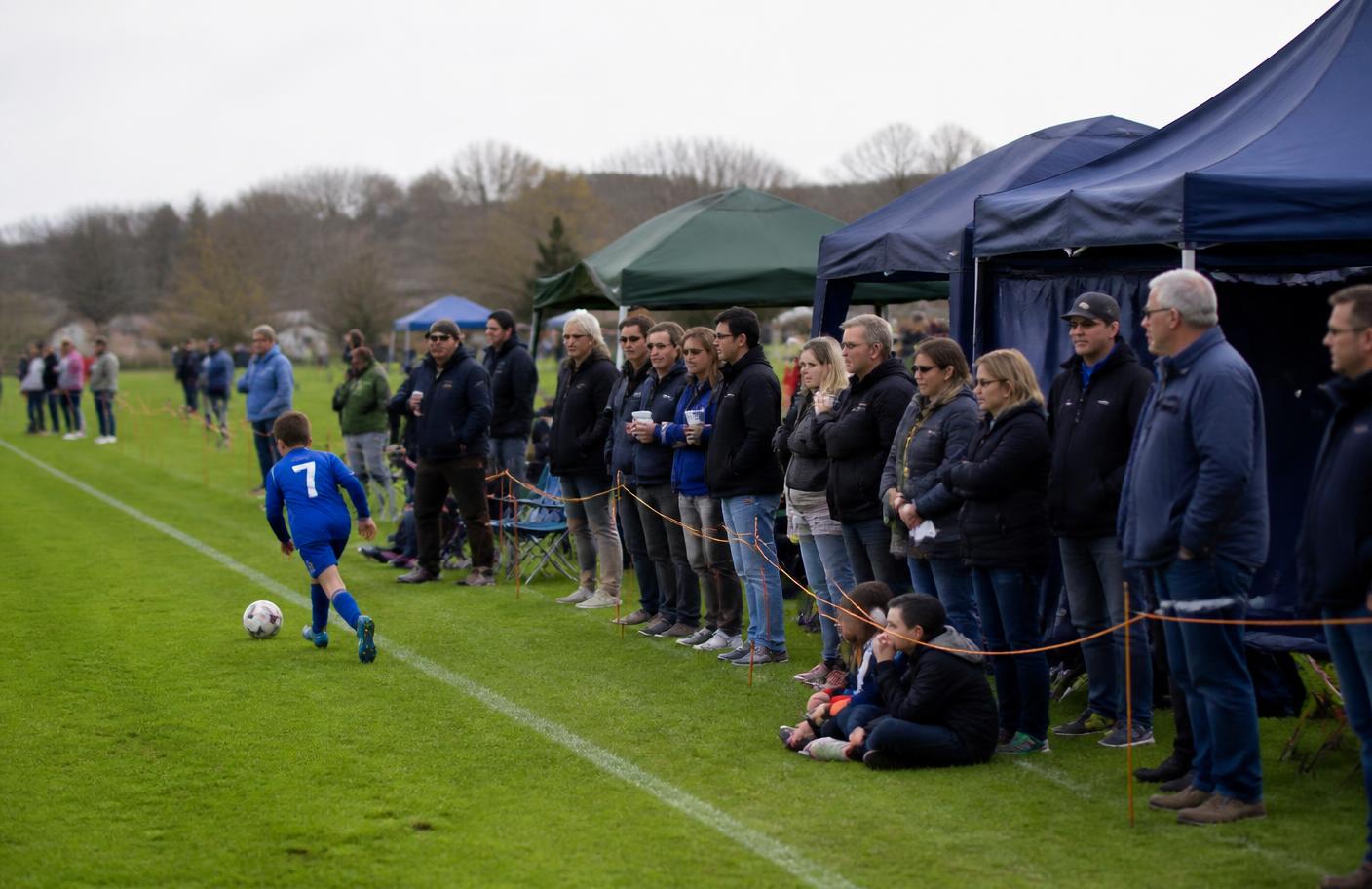 Spectators watching a junior football match on a grass pitch beside event gazebos